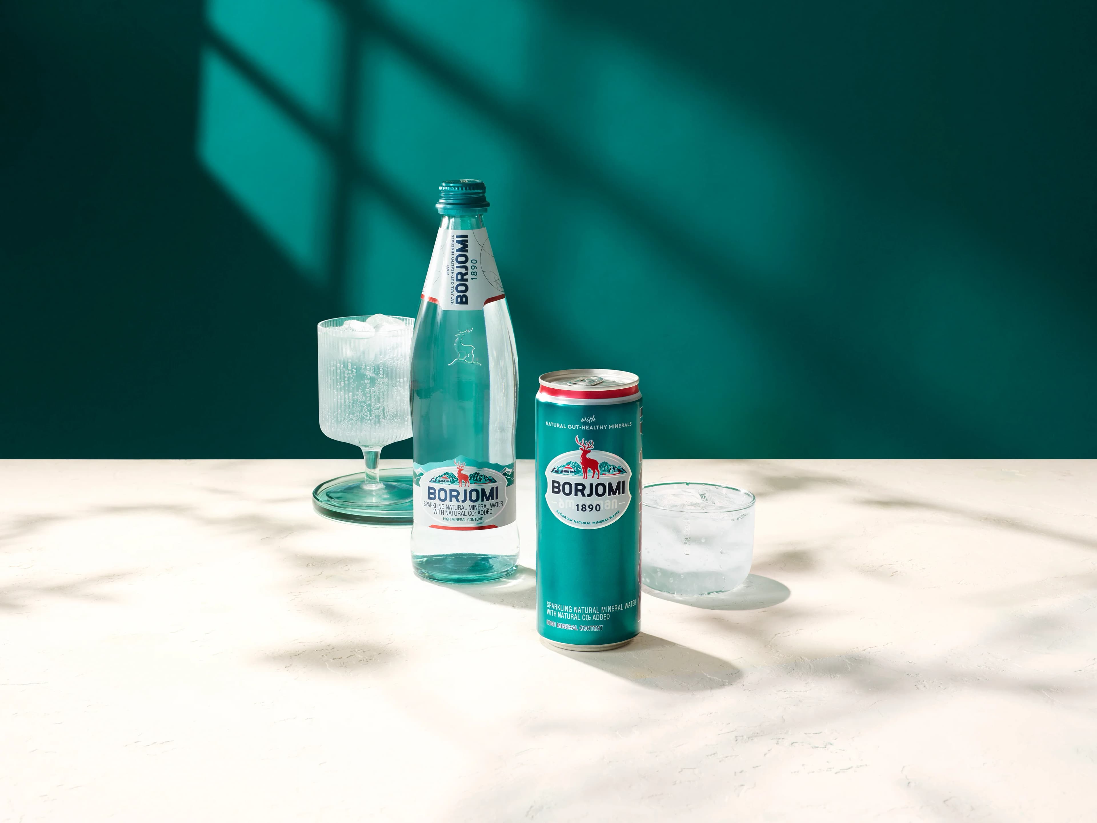 Borjomi glass bottle and can with ice water glasses on a marble table, teal background