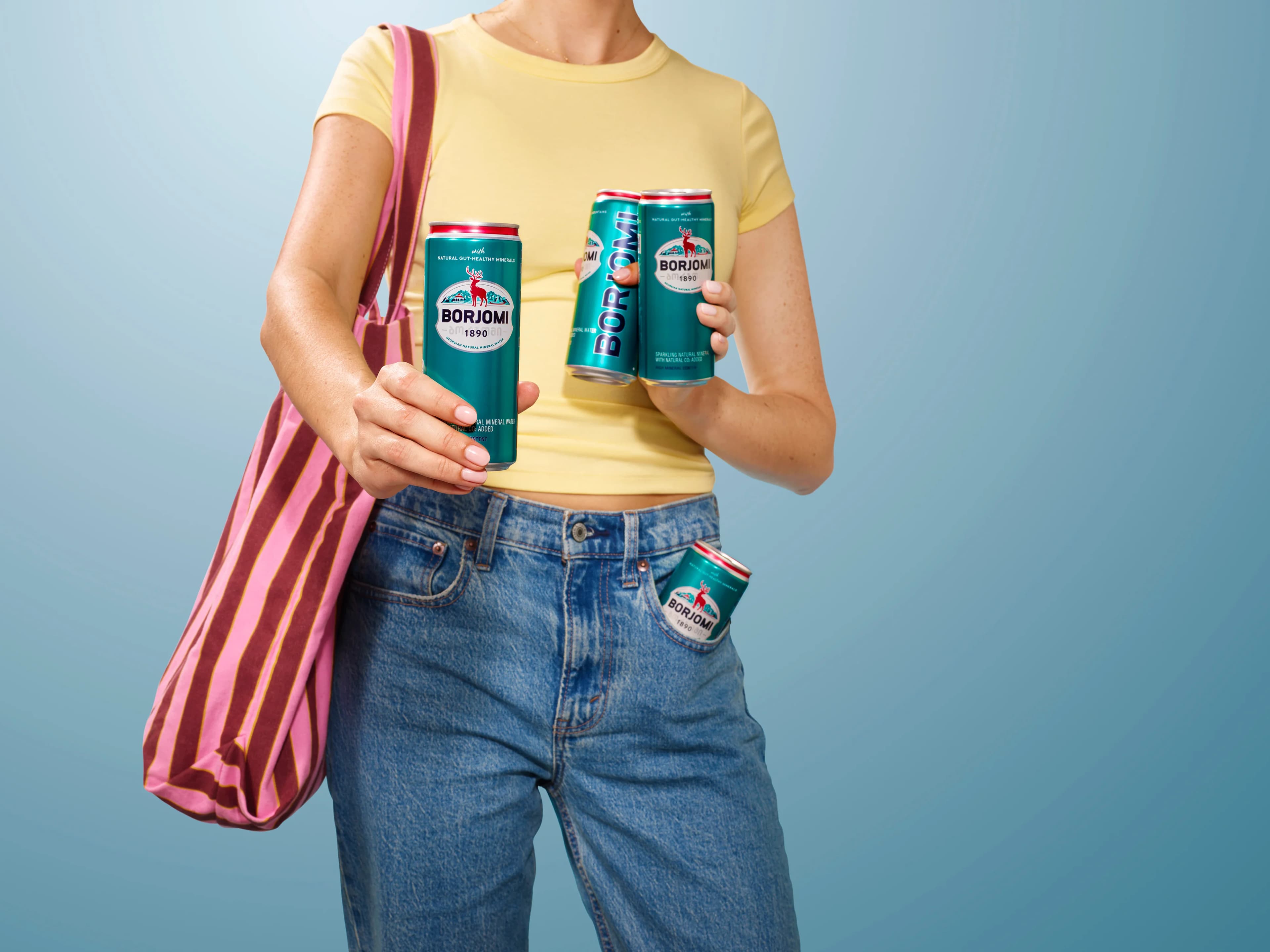 Woman holding multiple Borjomi cans including one in her pocket, light blue background