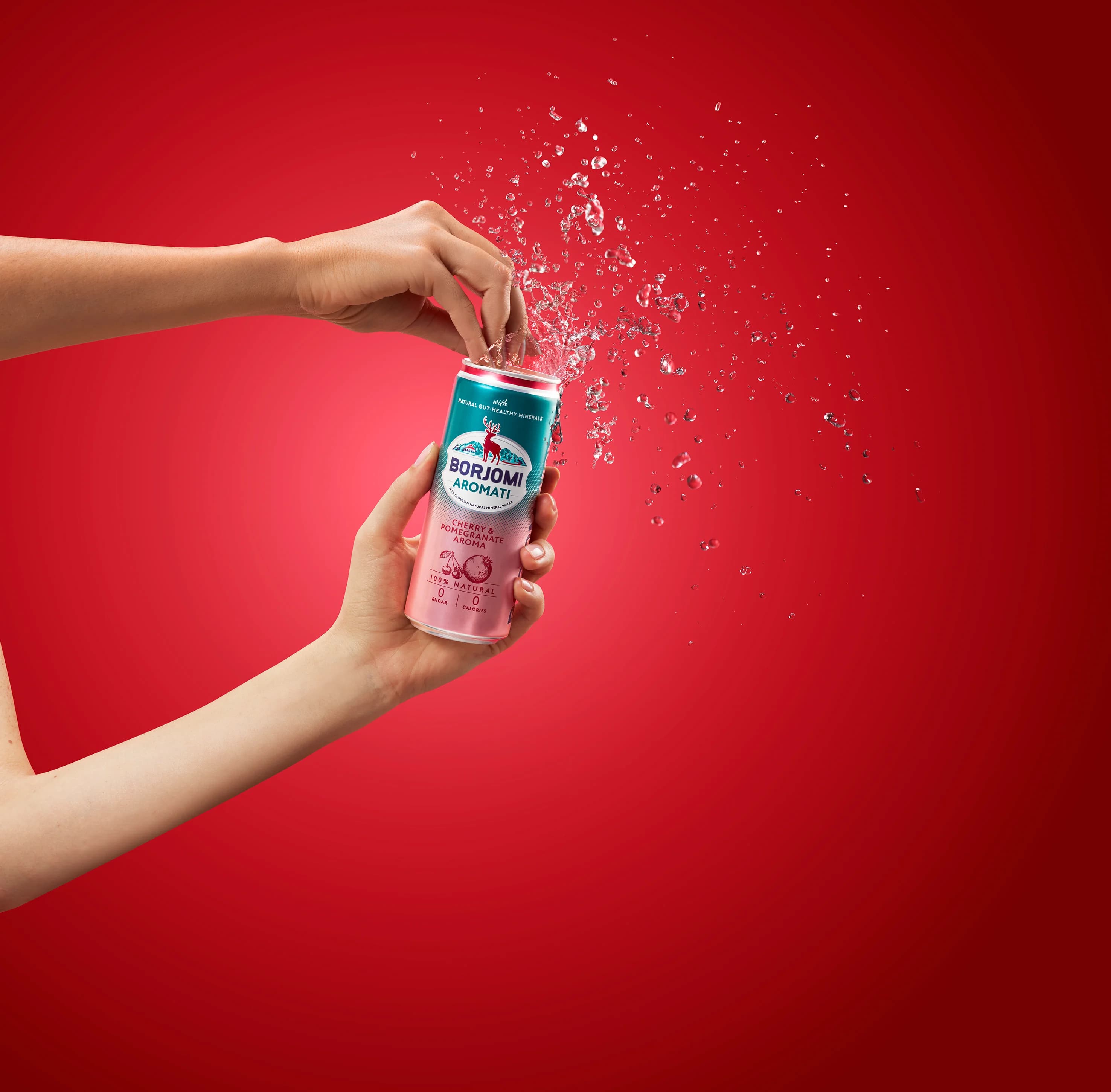Hands opening a Borjomi Aromati cherry pomegranate can with water splashing on a red background